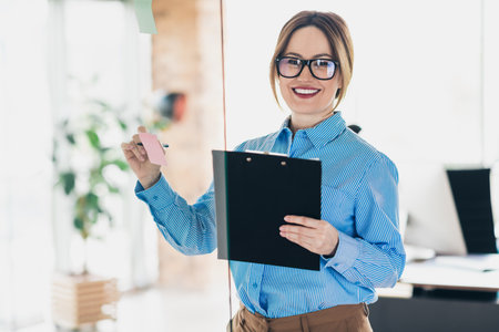 Confident businesswoman standing in office while planning and strategizing for projects, holding...
