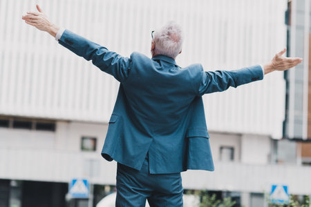 Rear view of a confident senior businessman standing outdoors in an urban setting with arms open wide facing the cityの写真素材