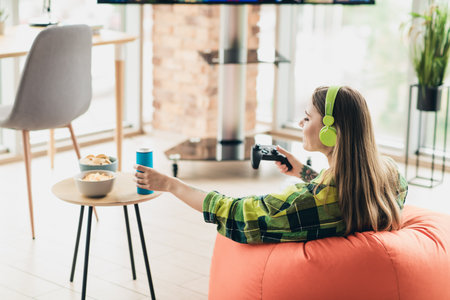 Cheerful woman relaxing indoors, playing video games and enjoying snacks and drinks while wearing...