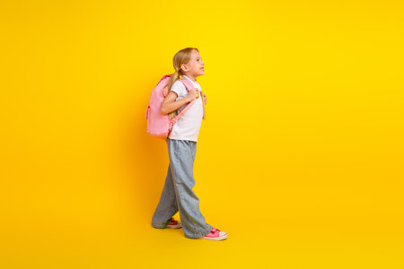 Adorable young schoolgirl with backpack walking against a yellow background - education, learning,...