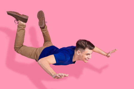 Cheerful young man pretending to fly against a pink background in casual attire, showcasing a fun...