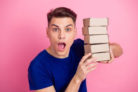 Excited young man holding a stack of carton boxes against a bright pink background in a trendy blue...