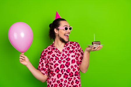 Young man celebrating with pink balloon and cake slice wearing festive party hat and sunglass...
