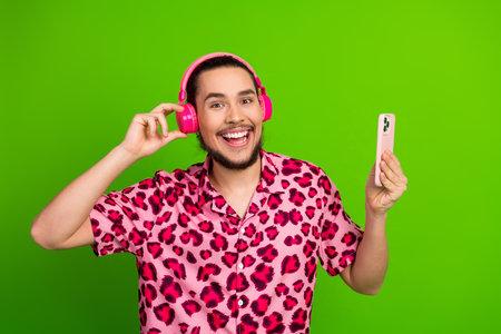 Young man with cheerful expression using pink headphones and holding a smartphone against green...
