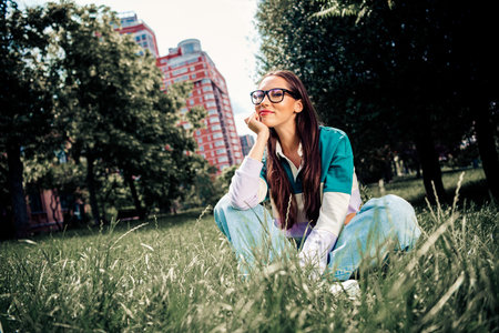Young woman enjoying leisure outdoors wearing casual attire sitting on grass against urban buildings...