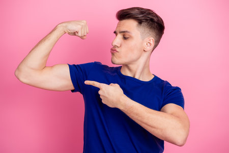 Confident young man in casual blue t-shirt flexing arm showing muscle on a pink background expressing pride and humorの写真素材