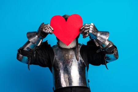 Medieval knight holding a red heart symbol on a blue background expressing love and bravery in a historically styled concept photograph.の写真素材