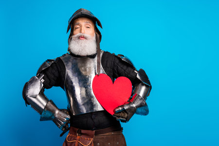 Senior man in metal armor holding heart symbol with vibrant blue background highlighting medieval themeの写真素材