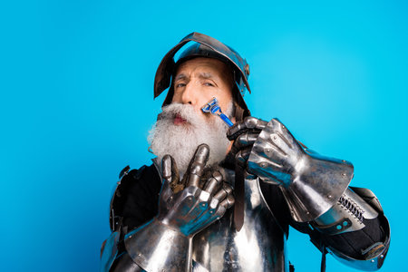 Elder knight in medieval armor whimsically shaving against blue background, showcasing humor and historical charm.の写真素材