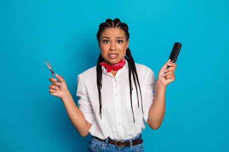 Confused young woman holding scissors and brush with expressive face on vivid blue backgroundの写真素材