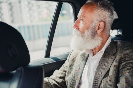 Mature businessman with a grey beard in a formal jacket looking out of a car window in an urban cityscape in daylightの写真素材