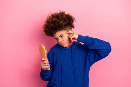 Young boy in blue hoodie holding a brush and pondering over grooming choices against a pink studio backgroundの写真素材