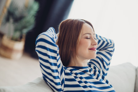 Relaxed young woman with closed eyes enjoying her time at home in a striped sweater on a bright sunny dayの写真素材