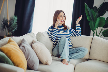 Young woman enjoying a phone call in a cozy living room with vibrant decor and stylish casual attire, relaxed at homeの写真素材