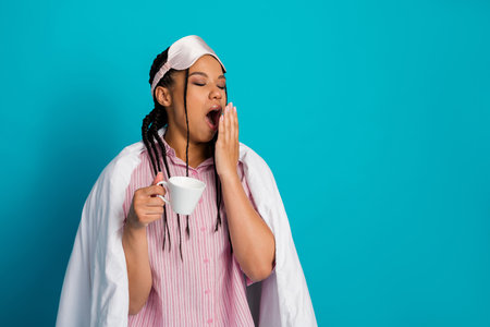 Tired young woman in pink pajamas yawning while holding a white cup against a bright blue backgroundの写真素材