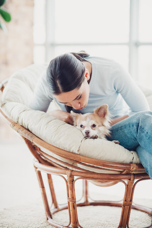 Beautiful woman in a cozy chair with her dog, sharing tender moments at home on a bright day.の写真素材
