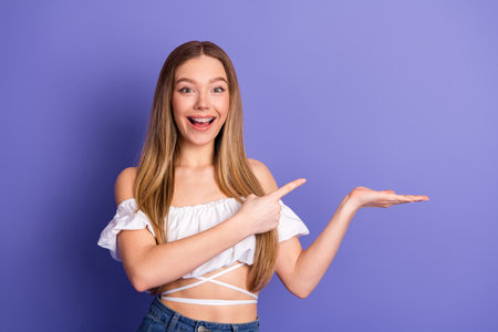 Excited young woman in casual white top and jeans points to promotional space against a purple background for advertising.の写真素材