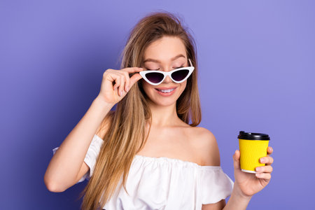 Trendy young woman holding a yellow coffee cup against a purple background, showcasing her chic style and joyful expressionの写真素材