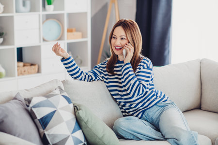 Young woman in striped sweatshirt enjoying conversation on phone at home in stylish living room interiorの写真素材