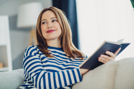 Charming young woman enjoying leisure at home while reading a book, sitting on a cozy couch in natural daylightの写真素材