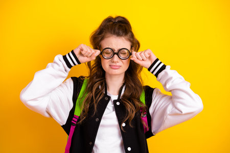 Cheerful young student with glasses rubbing eyes in a vibrant yellow background showing school spirit and learningの写真素材