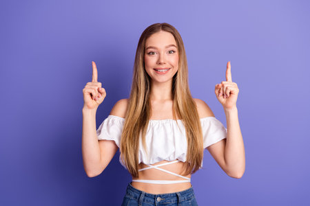 Happy young woman pointing upwards on a vibrant purple background, conveying a promotional concept with a trendy casual lookの写真素材