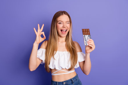 Lovely Smiling young woman holding chocolate bar and making gesture against violet backgroundの写真素材