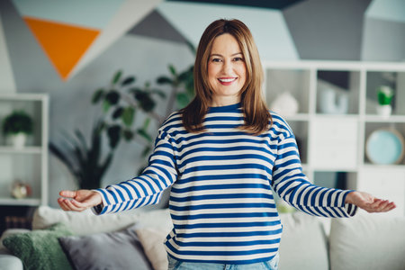 Charming woman wearing a striped sweatshirt posing happily in a stylish living room with modern and vibrant decorの写真素材