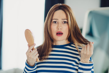 Worried woman in a striped casual outfit at home holding a hairbrush and examining her hair loss, concerned expressionの写真素材