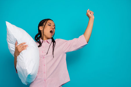 Young woman in pink pajamas stretches energetically holding a pillow, conveying cheerful morning vibes against a blue backdropの写真素材