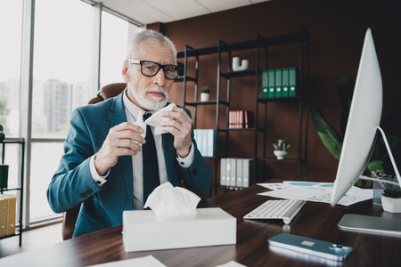 Mature business professional with a tissue, working at office desk, portraying professionalism in a corporate environmentの写真素材