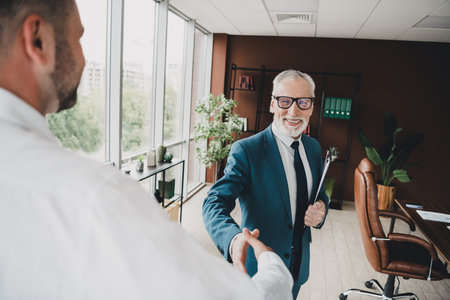 Confident senior businessman shakes hands in a professional office environment, symbolizing partnership and career successの写真素材