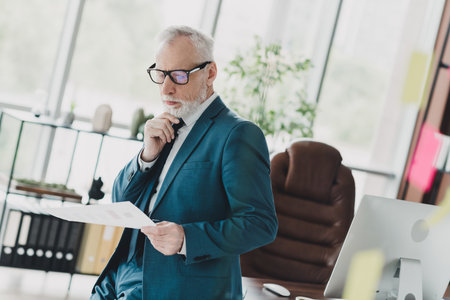 Senior businessman reviewing documents thoughtfully while standing in a modern office environment with a confident demeanorの写真素材