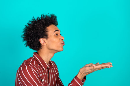 Funny man with curly hair kissing his hand playfully in a joyful gesture against a vibrant turquoise...