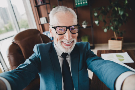 Smiling senior businessman in a formal suit taking a selfie in a modern office space with professional settingの写真素材