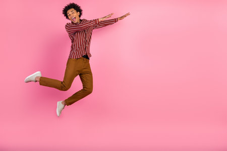 Young carefree man jumping joyfully against a vibrant pink background in trendy attire, spreading...