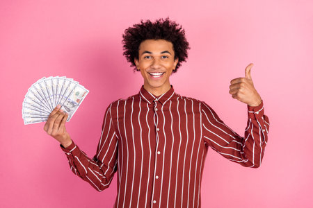 Young man in striped shirt holding cash and giving thumbs-up against pink background, expressing joy...