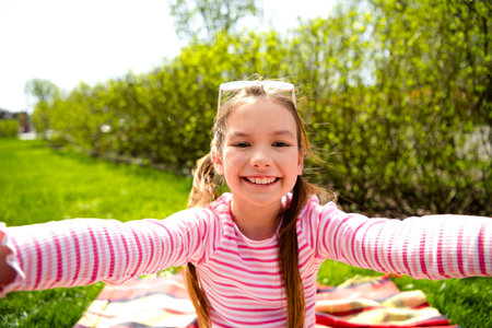 Smiling young girl enjoying a sunny day outdoors while sitting on a blanket in a park surrounded by...