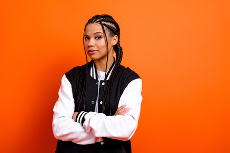 Confident young woman with braided hairstyle and varsity jacket standing against vibrant orange...