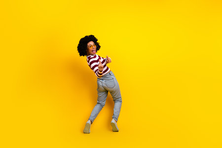 Funny woman with curly hair in casual striped top and jeans posing joyfully against a bright yellow backgroundの写真素材