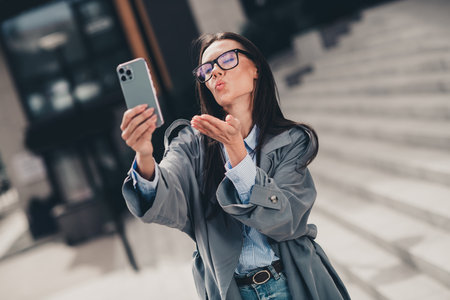 Elegant young woman taking a selfie outdoors in urban setting wearing a stylish trench coat during daylight hoursの写真素材