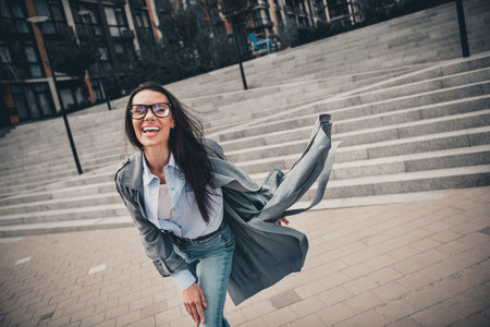 Elegant young woman wearing a stylish gray trench coat outdoors in an urban environment on a sunny dayの写真素材