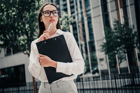 Successful young woman holding a clipboard, contemplating ideas outdoors in a modern urban setting surrounded by high-rise buildingsの写真素材
