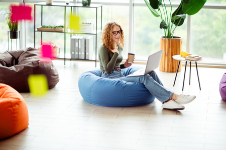 Young woman enjoys a relaxing workspace in contemporary office setting with laptop and coffee, sitting on a beanbagの写真素材