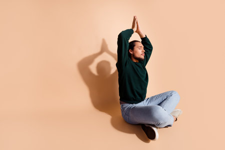 Young man sitting cross-legged on beige background meditating in green sweater, embracing mindfulness and relaxationの写真素材