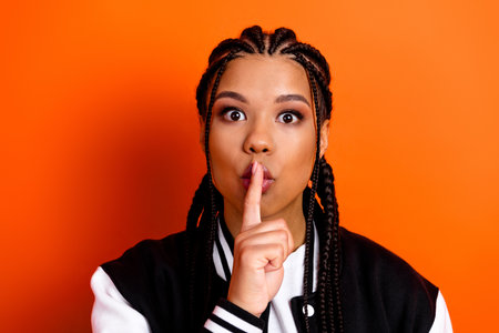 Portrait of a young woman with braided hair making a quiet gesture against vibrant orange background