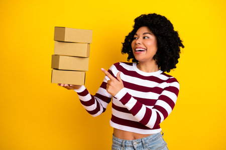 Smiling young woman holding stacked cardboard boxes in front of a vibrant yellow background...