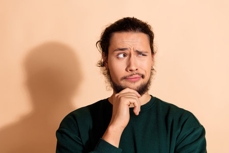 Thoughtful young man in casual fashion posing on beige background with a curious expression and...