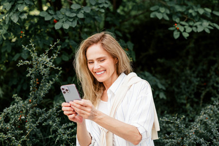 Charming middle-aged woman using smartphone outdoors enjoying sunny day in park surrounded by...