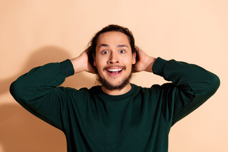 Young male with a cheerful expression against a beige background, wearing green sweatshirt, hands on...